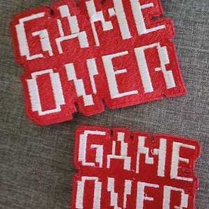 May include: Two red embroidered patches with the words "GAME OVER" in white, pixelated font. The patches are rectangular with a slightly distressed edge, and are set against a gray background.