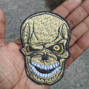 May include: Embroidered patch featuring a stylized skull with a menacing expression. The skull has a tan and black color scheme, with green and red eyes and a wide, toothy grin. The patch is likely designed for application on clothing or accessories.