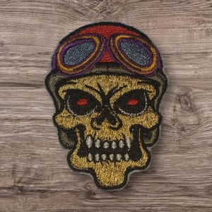 May include: Embroidered patch featuring a skull wearing a red and purple aviator helmet with goggles. The skull is gold with black details and red eyes. The patch is likely for jackets, bags, or other apparel.
