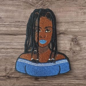 May include: Embroidered patch featuring a woman with dark brown skin, black braided hair, and blue lips. She is wearing a blue off-the-shoulder top.