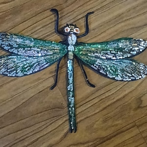 May include: A large, decorative dragonfly applique with iridescent green and silver sequins on its wings. The dragonfly has a black body and antennae, with orange and black accents on its head. The applique is designed to be sewn onto fabric.