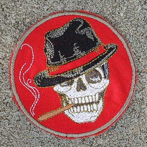 May include: Embroidered patch featuring a skull wearing a black fedora and smoking a cigar. The skull is white with detailed teeth, set against a red circular background with a gray border. A wisp of smoke curls upwards.
