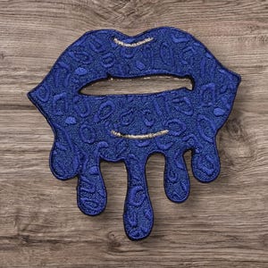 May include: A blue, dripping, embroidered lip patch with a gold outline. The patch is made of a textured fabric and has a slightly raised design.