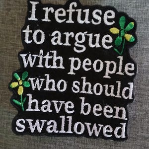 May include: Black embroidered patch with the text "I refuse to argue with people who should have been swallowed." The patch has white lettering and two small flower designs with green and yellow petals.