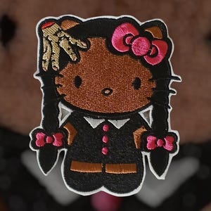 May include: Embroidered patch featuring a cartoon character with dark skin, black hair in pigtails, and a pink bow. The character wears a black outfit with a white collar and a gold hand gesture. The patch has a white border.