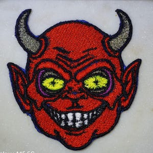 May include: Red embroidered patch of a devil's face with yellow eyes and a toothy grin.