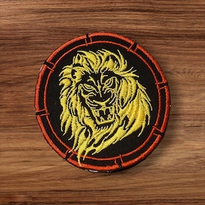 May include: Round embroidered patch featuring a yellow lion's head on a black background. The lion has a fierce expression, with a detailed mane and open mouth. The patch is bordered by a red ring with stitched details.