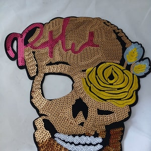 Puede incluir: Una calavera de lentejuelas doradas con un diseño rosa de "R.I.P." en la frente, una rosa amarilla en el ojo derecho y flores azules en el lado derecho.