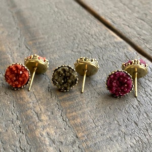 May include: Three pairs of stud earrings with a druzy-like appearance. The earrings have a gold-colored crown-shaped setting. The druzy stones are in shades of red, olive green, and burgundy.