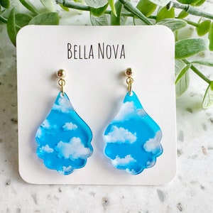 May include: A pair of blue and white cloud earrings with gold hooks. The earrings are shaped like teardrops and have a scalloped edge. The earrings are labeled "Bella Nova".