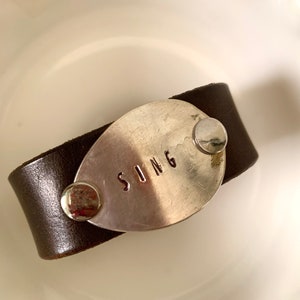 Op de afbeelding: Een donkerbruine leren armband met een zilverkleurig metalen accent. Het metalen stuk is ovaal van vorm en gestempeld met het woord "SONG". De armband is vastgezet met zilveren klinknagels.