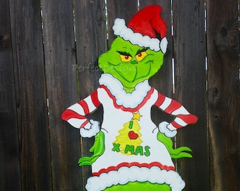 Op de afbeelding: Een groen Grinch-personage dat een rood-wit gestreept shirt draagt met een kerstboomgrafiek en de woorden "I  XMAS" erop. De Grinch draagt een kerstmanmuts en heeft een ondeugende uitdrukking op zijn gezicht.