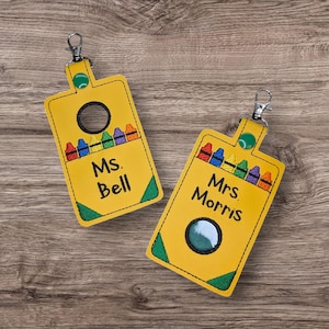 May include: Two yellow keychains shaped like crayon boxes, each with a silver clasp. One keychain says "Ms. Bell" and the other says "Mrs. Morris". Both have a colorful crayon design and a circular opening.