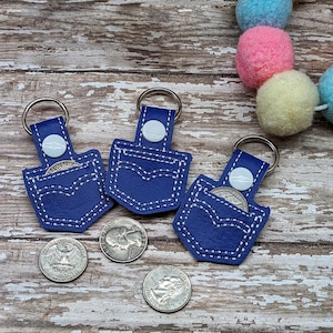 Könnte beinhalten: Drei blaue Leder-Schlüsselanhänger mit einem gestickten Taschen-Design. Jeder Schlüsselanhänger hat einen silbernen Ring und einen weißen Kreis in der Mitte der Tasche. Die Schlüsselanhänger befinden sich auf einer Holzoberfläche mit vier bunten Kugeln im Hintergrund.