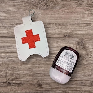 May include: White hand sanitizer holder with a red cross embroidered on it. The holder has a silver clip and is attached to a small bottle of hand sanitizer.