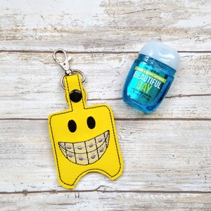 May include: Yellow felt keychain with a smiling face and braces, holding a blue plastic bottle of Bath & Body Works "Beautiful Day" antibacterial hand gel. The bottle is 1 fl oz / 29 ml.