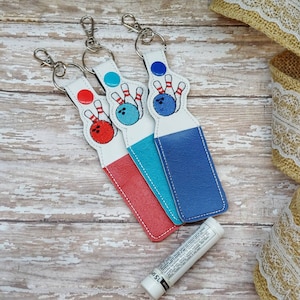 以下が含まれることがあります： ボウリングボールとピンの刺繍デザインが施されたキーホルダーが3つ。キーホルダーは赤、青、ターコイズブルーのファブリックホルダーに取り付けられています。
