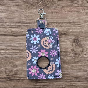 Peut inclure: Un porte-flacon de désinfectant pour les mains à imprimé floral gris avec un trou noir pour le flacon. Le support présente un motif de fleurs souriantes, de citrouilles et d'étoiles roses, pêche et bleues. Il a un clip argenté et une fermeture à bouton-pression noire.