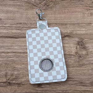 May include: A light grey and white chequered pattern keychain holder, with a silver-tone clasp and a grey button closure. The holder has a circular opening near the bottom.