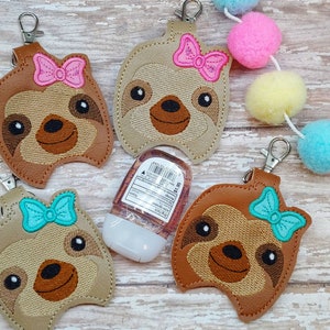 May include: A collection of sloth-shaped keychains in various shades of brown and tan, each adorned with a colorful bow. The keychains have a metal clasp for easy attachment. A small bottle of hand sanitizer is also present.