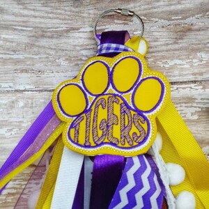 May include: Purple, white, and yellow ribbon keychain with a yellow paw print with the word "TIGERS" embroidered in purple.