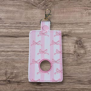 May include: A rectangular keychain holder featuring a repeating pattern of light pink bows on a white and light pink vertical striped background. The holder includes a circular cutout with pink stitching and a gold-toned metal lobster clasp.