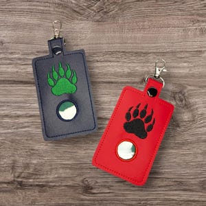 May include: Two keychains with a rectangular design. One is navy blue with a green paw print, and the other is red with a black paw print. Each keychain has a silver-colored clip and a circular element.