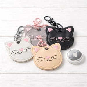 Könnte beinhalten: Vier katzenförmige Schlüsselanhänger aus weißem, grauem, schwarzem und beigem Leder. Jeder Schlüsselanhänger hat rosa Akzente an Ohren und Nase sowie gestickte Schnurrhaare. Ein silbernes Apple AirTag liegt neben den Schlüsselanhängern.