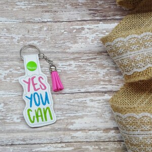 May include: White keychain with a pink tassel. The keychain has the words "YES YOU CAN" embroidered in blue, green, and yellow.