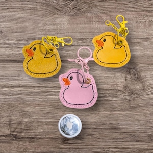 May include: Three duck-shaped keychains in yellow and pink, with orange beaks, and a silver AirTag. The keychains have yellow and pink clasps. The duck keychains are on a wooden surface.