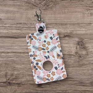 May include: A floral print waste bag dispenser with a silver-colored clip. The dispenser features a white background with pink, brown, and gray floral patterns. The dispenser has a circular opening with a pink border.