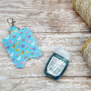 May include: A blue and white hand sanitizer holder with a keychain and a flamingo, donut, and inflatable pool float pattern. The holder is attached to a small bottle of hand sanitizer.