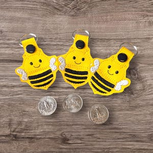 Puede incluir: Tres llaveros amarillos con forma de abeja, con rayas negras y caras sonrientes. Cada llavero tiene un llavero plateado y un botón negro. Tres monedas de plata están debajo. Sobre una superficie de madera.