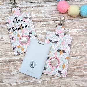 May include: Two keychains with a geometric pattern in pink, blue, and gold. One keychain has a clear window and says "Mrs. Bobbie". The other keychain has a clear window and a gray card with a logo that says "CENTEGIX".
