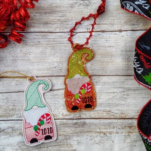 May include: Two Christmas ornaments shaped like gnomes with red hats, white beards, and green hats. The gnomes are holding candy canes and have the year 2020 embroidered on their shirts.