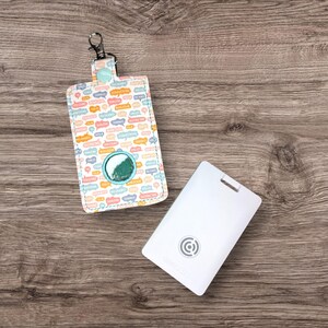 Puede incluir: Una bolsa blanca y de color pastel con una ventana transparente y una pequeña etiqueta rectangular blanca con un logotipo circular. La bolsa tiene un patrón colorido de burbujas de diálogo con palabras en diferentes idiomas. La bolsa tiene un clip plateado.