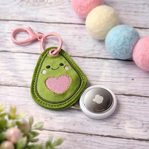 May include: A green avocado-shaped keyring with a pink heart and smiling face embroidery. The keyring has a pink clasp and is next to a white AirTag. Pastel-coloured felt balls are in the background.