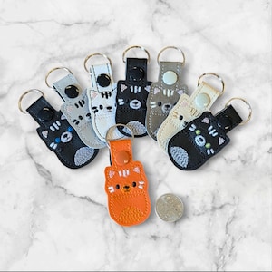 Puede incluir: Un conjunto de ocho llaveros con diferentes gatos de colores con caras bordadas. Los llaveros están hechos de cuero y tienen un anillo de metal plateado.