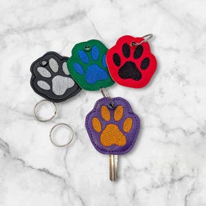 May include: Four colorful paw print keychains. The keychains are in the shape of a paw print, with a silver key ring. The colors are black with white, green with blue, red with black, and purple with orange.