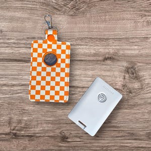 Puede incluir: Un porta bolsas para excrementos de perro a cuadros blancos y naranjas con un clip plateado y una tarjeta de plástico blanca con el texto "Agility" impreso en ella.