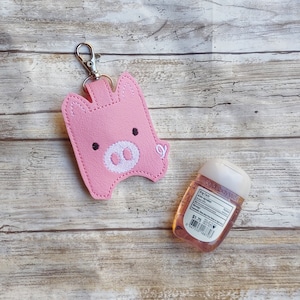 May include: Pink felt pig keychain with a silver key ring. The pig has a white snout and black eyes. A small bottle of hand sanitizer is also in the image.