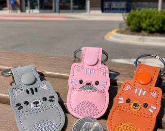 May include: Three cat-shaped keychains in grey, pink, and orange leather-like material, each featuring a snap button, a metal ring, and a small pocket containing a US quarter coin for scale, showing the coin's diameter of approximately 2.4 cm.