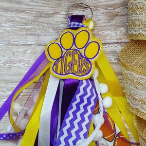 May include: A yellow and purple paw print keychain with the word "TIGERS" embroidered on it. The keychain has a variety of ribbons and bows attached to it, including yellow, purple, white, and a chevron pattern.