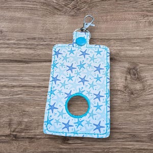 May include: A light blue fabric bag with a starfish pattern in shades of blue. The bag has a circular opening and a silver-colored clip. The bag is designed to hold a roll of something, such as dog waste bags.