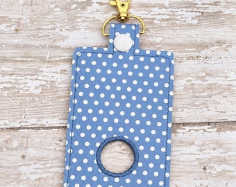 May include: A rectangular blue badge holder featuring a white polka dot pattern, a circular cutout in the center with dark blue stitching, a white snap button closure at the top, and a gold-toned metal lobster clasp attached to a D-ring.