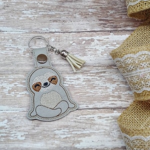 May include: A gray sloth keychain with a smiling face and brown eyes. The keychain has a silver ring and a tassel.