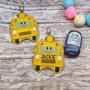 May include: Two yellow school bus-shaped hand sanitizer holders with smiling faces. The holder on the right has the name "JACKIE" embroidered on it.