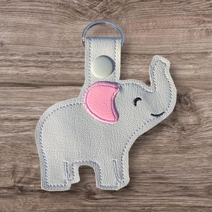 May include: A light gray elephant-shaped keychain with a pink ear and a silver key ring. The elephant has a smiling face and is made of a textured material. The keychain is attached to a light gray tab with a white button.
