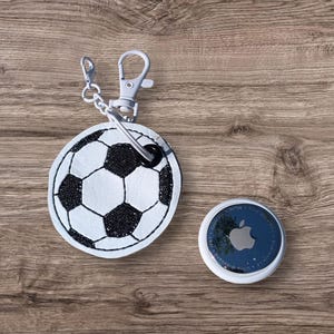 May include: A white and black soccer ball keychain with a silver clasp and chain next to a white and blue Apple AirTag. The soccer ball design is stitched onto a round leather piece.