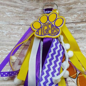 May include: A yellow and purple paw print keychain with the word "TIGERS" embroidered on it. The keychain has multiple ribbons in yellow, white, and purple, with white pom-poms attached.
